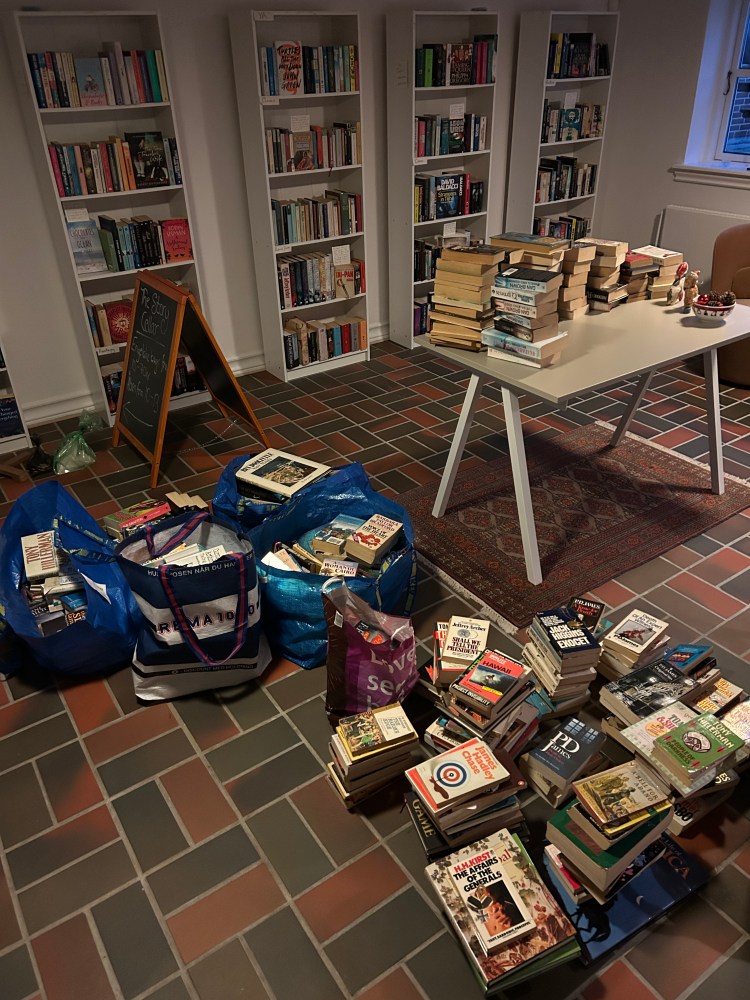 Books stacked on the floor. Books in IKEA bags. Books stack on a table in front of full bookshelves. 